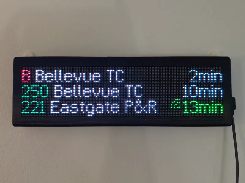 An RGB LED matrix display mounted in a frame hanging on a wall. It reads a list of three different public transit routes, their destinations, and the estimated arrival time for each.